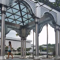 Kasia Porebska caminando bajo estructura islámica entre columnas decorativas y vegetación tropical